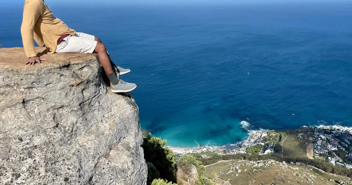 Las piernas de un senderista colgando sobre el borde de una roca, muy por encima de un mar azul intenso