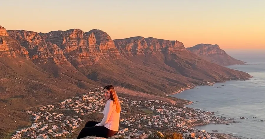 Eine Frau sitzt bei Sonnenuntergang auf einem Felsen über Camps Bay und den Twelve Apostles